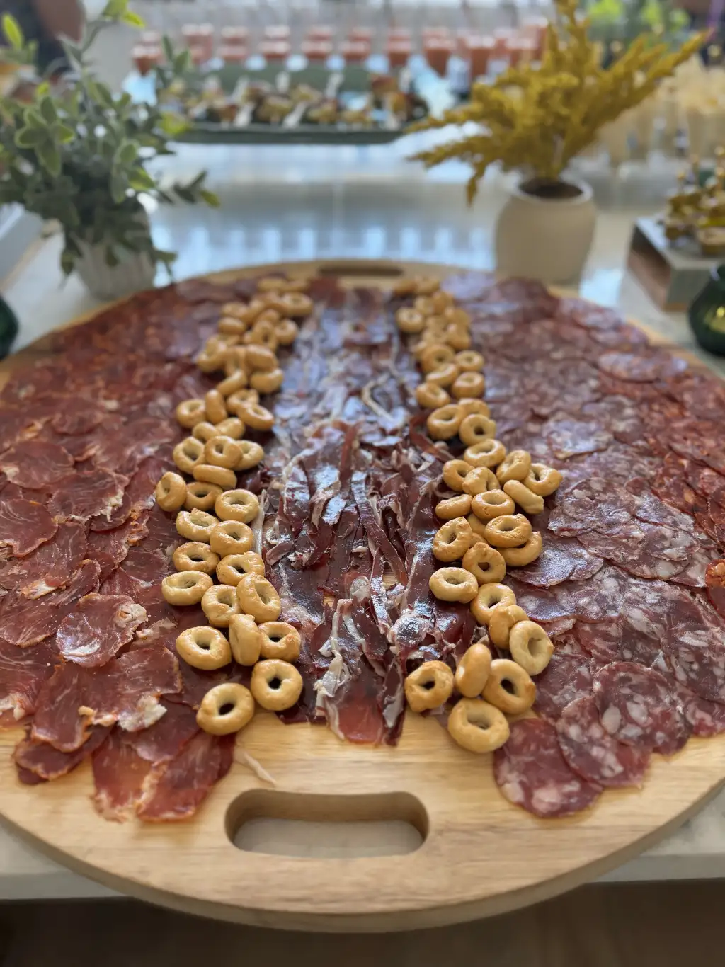 Round Spanish charcuterie board with three cured meats and picos crackers, set in a private-home dining room with greenery — Cantábrico, Houston